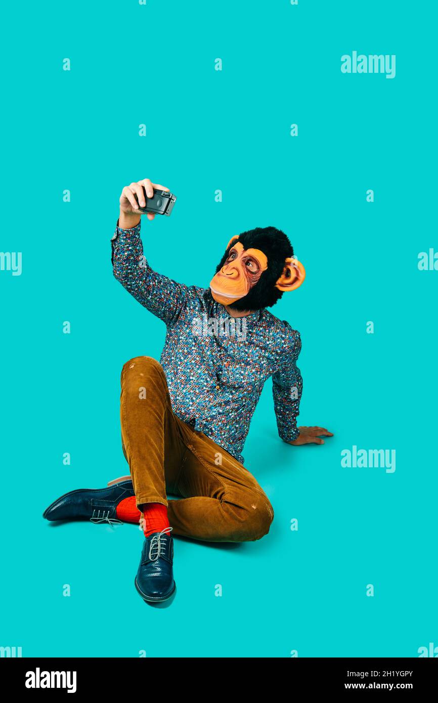 a young man, wearing a monkey mask, takes a selfie with a retro filma camera, sitting on a blue background with some blank space on top Stock Photo