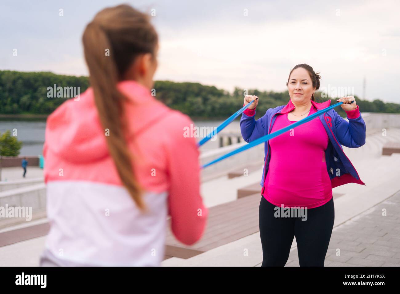 Close-up view from back of fat young woman doing exercises using resistance band for weight loss with personal trainer outdoor in summer day. Stock Photo