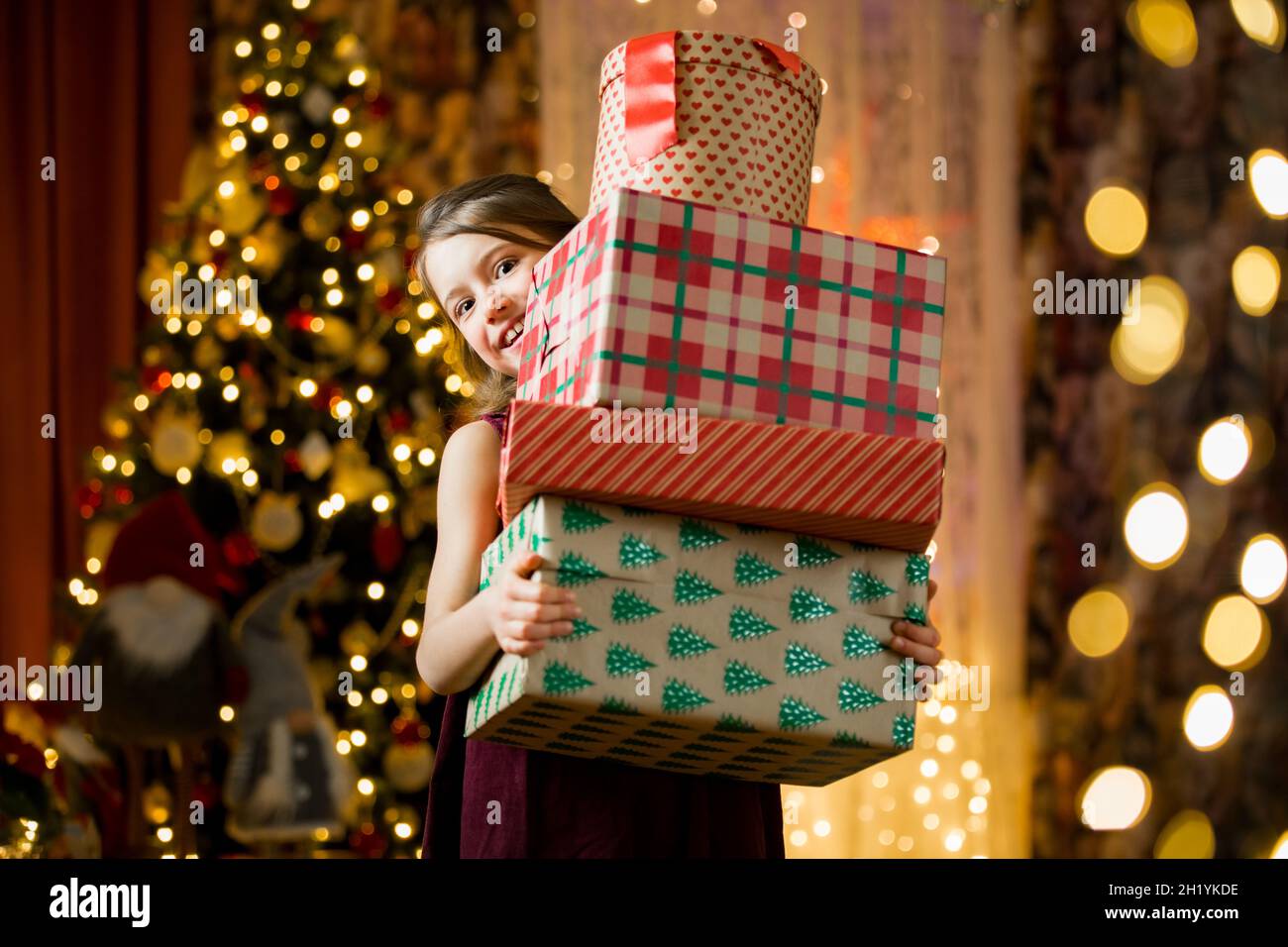 Excited curious little girl smiling, holding pile of Christmas gifts. Beautifully decorated loving room with lights and Christmas tree. Happy holiday Stock Photo