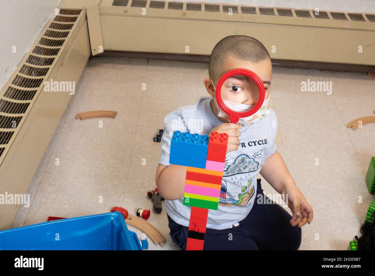 Education Preschool 3-4 year olds boy using magnifying glass to look at tower he built from colorful plastic connecting briks (Duplo) Stock Photo