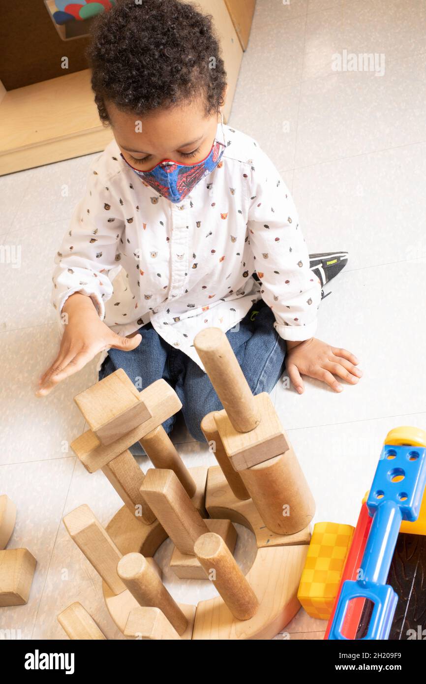 Education Preschool 3-4 year olds boy playing in block area putting wooden block on top of tower he built, wearing face mask to protect from Covid-19 Stock Photo