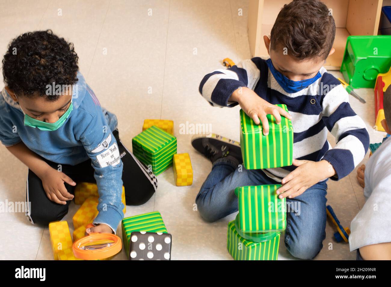 Education Preschool 3-4 year olds two boys playing separately in classroom, one building with blocks the other using a magniifying glass both wearing Stock Photo