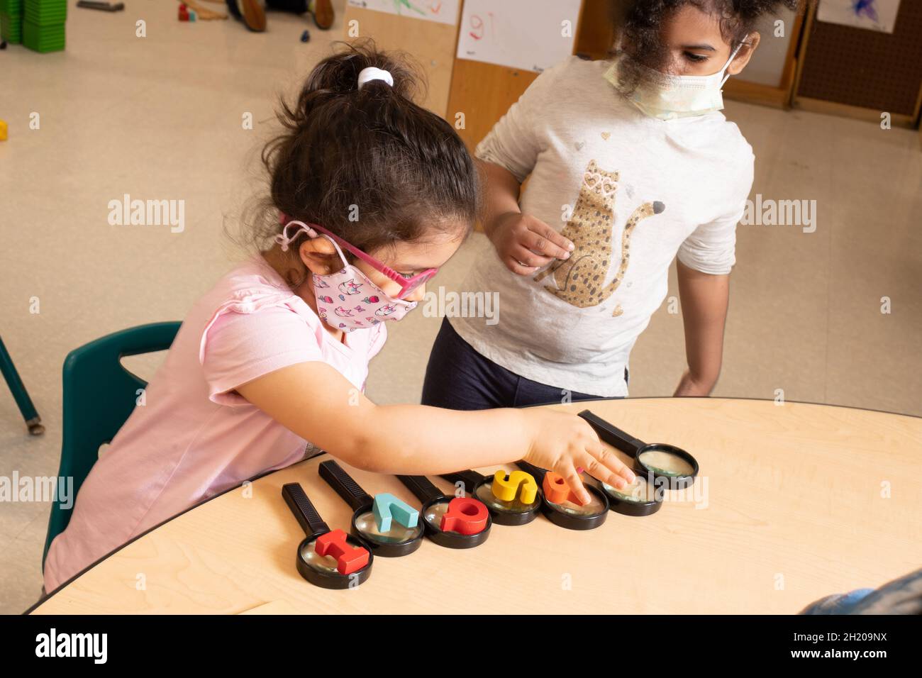 Education Preschool 3-4 year olds two girls playing with magnifying glasses, one girl putting wooden alaphabet letters on each magnifying glasses line Stock Photo