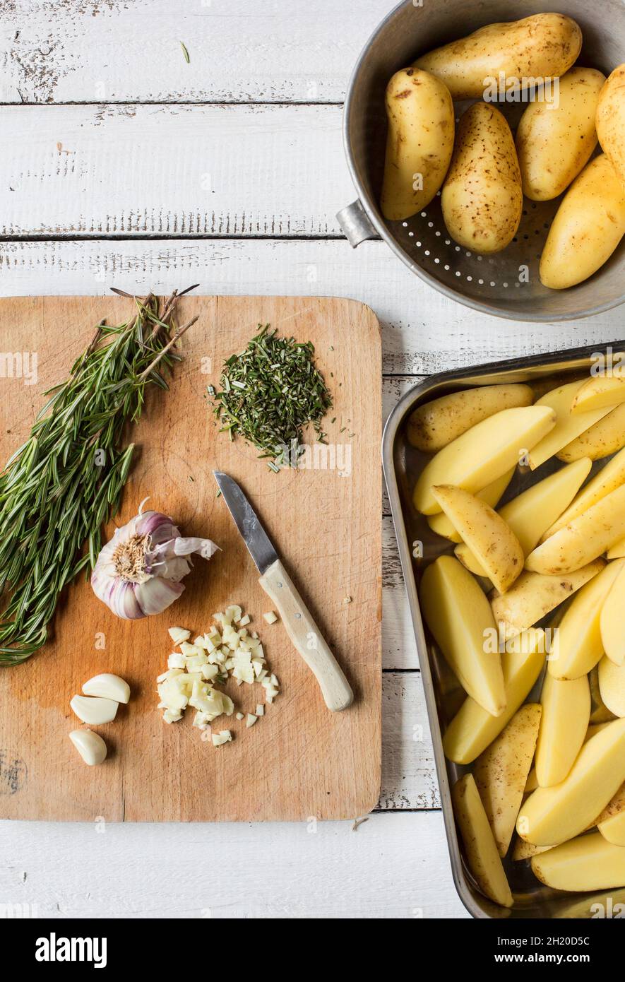 Ingredients for rosemary potatoes on a wooden chopping board and raw potato wedges in a roasting dish Stock Photo