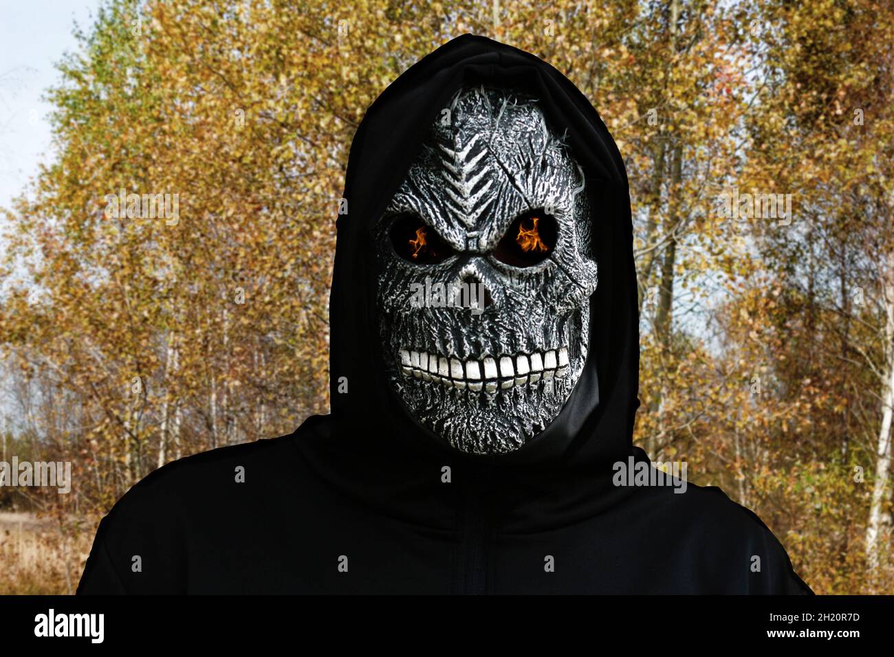 Close-up portrait of grim reaper. Man in death mask with fire flame in eyes on yellow aspen tree background. Carnival costume, creepy teeth. Halloween Stock Photo