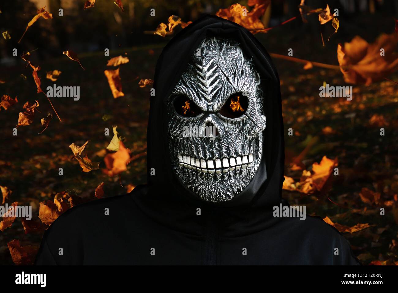Close-up portrait of grim reaper. Man in death mask with fire flame in eyes on magic autumn flying dry leaves background. Carnival costume, creepy Stock Photo