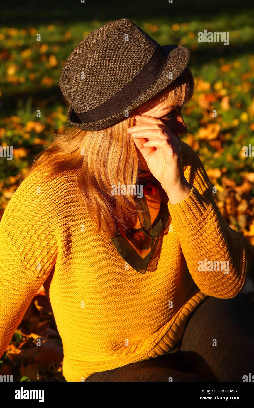Portrait of candid young Caucasian blonde woman in colorful autumn park. Bright stylish woman in yellow pullover, black hat and shawl of neck on fall Stock Photo