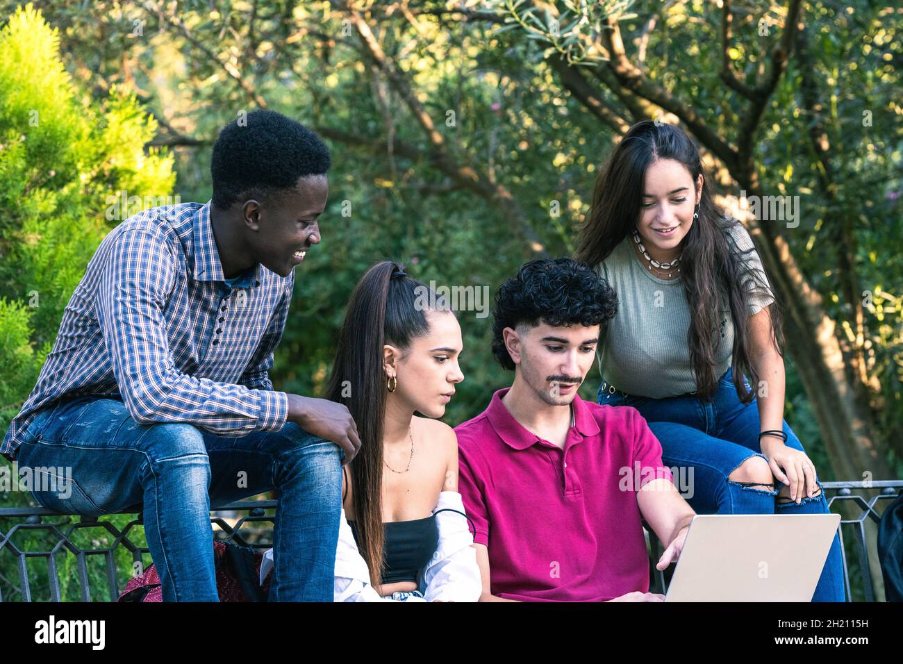 Four multiethnic friends looking at a laptop while sitting on a bench of a park Stock Photo