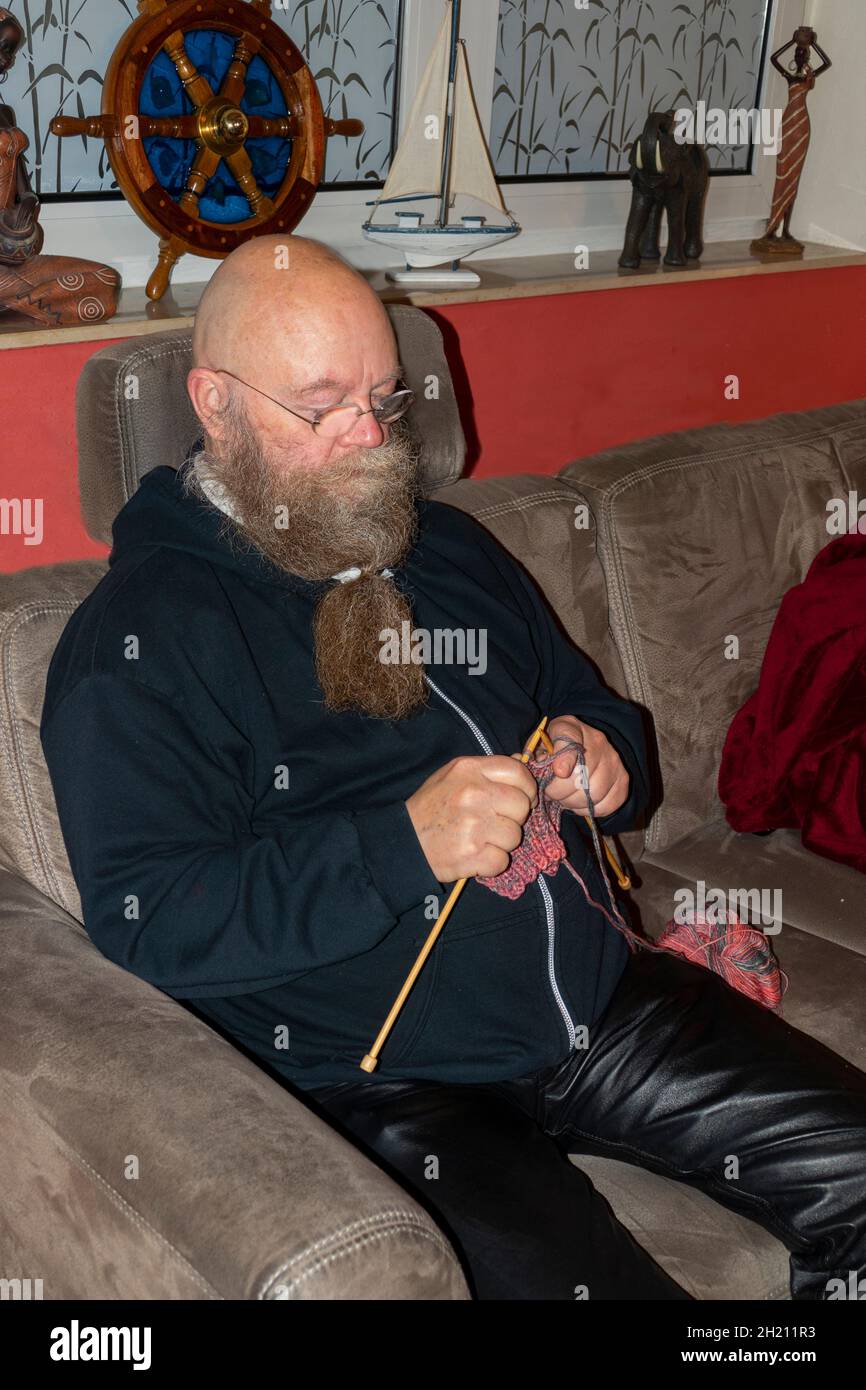 Mann mit langem Bart und Brille sitzt auf der Couch und strickt einen Schal Stock Photo