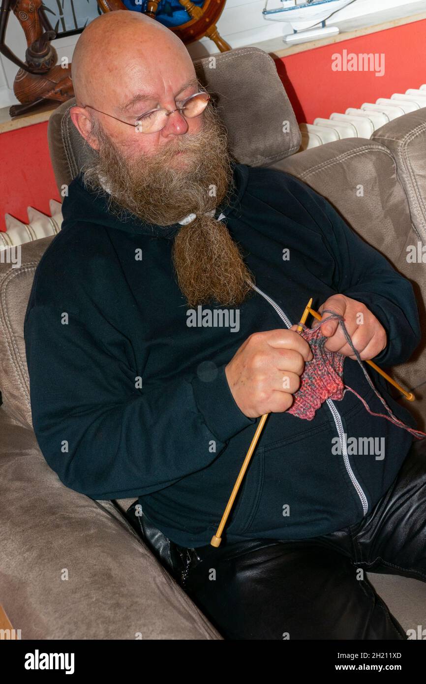 Mann mit langem Bart und Brille sitzt auf der Couch und strickt einen Schal Stock Photo