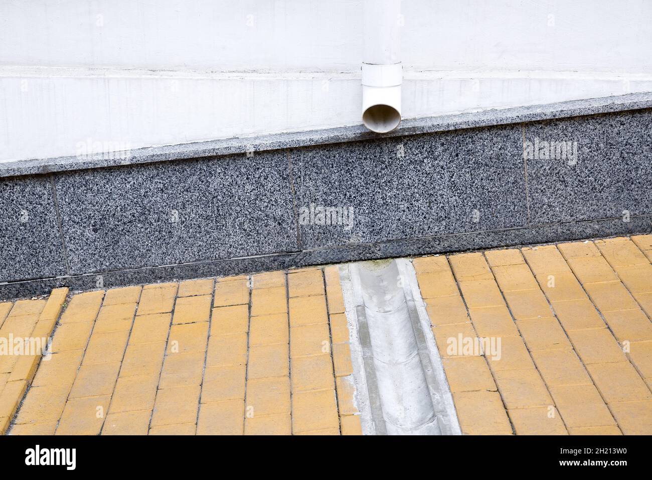 gray granite cladding on facade of a house wall with a drainage pipe and a concrete dish canal on a stone slab sidewalk to drain rainwater front view. Stock Photo