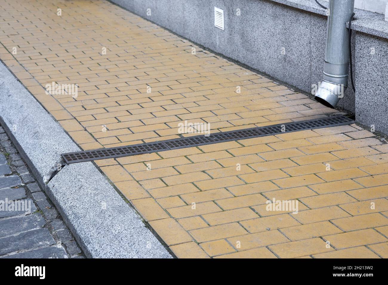facade of building with granite stone cladding with storm pipe into drainage canal with iron grating on pedestrian sidewalk made of stone tiles and cu Stock Photo
