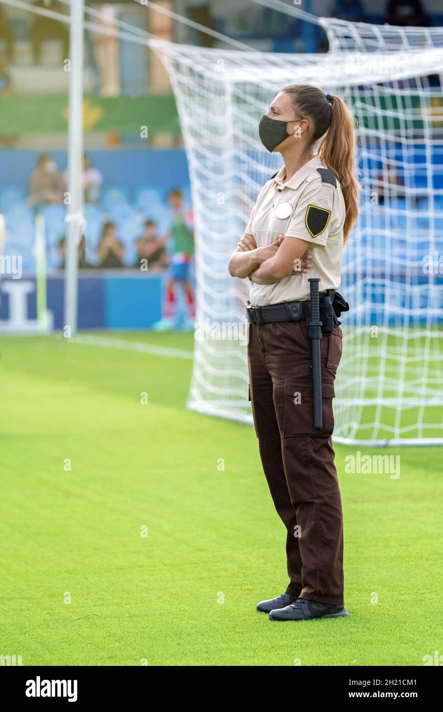 Woman standing at the side, uniformed as a security guard wearing a mask, stares straight ahead. Stock Photo