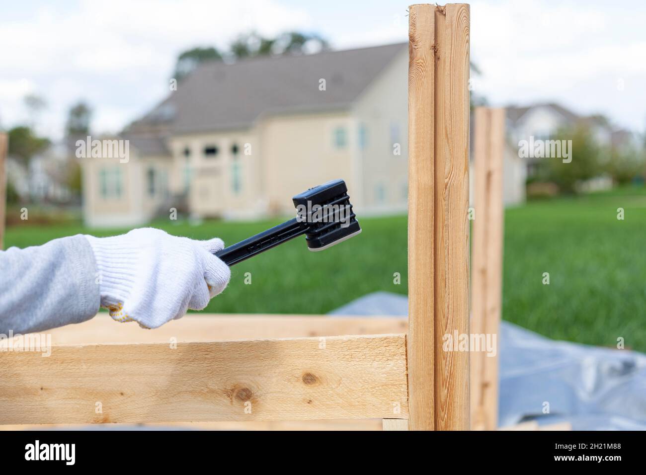 outdoor diy project in backyard with lawn. A person wearing safety gloves is installing a wooden fence or furniture using a plastic hammer to tighten. Stock Photo