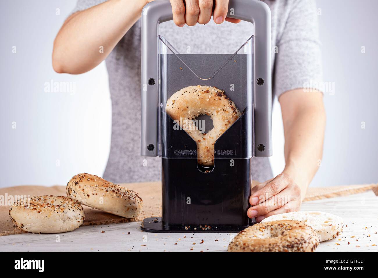 Cacuasian woman wearing short sleeve t shirt is slicing fresh baked bagels using manual bagel slicer. She places the bagel inside the holder and puts Stock Photo
