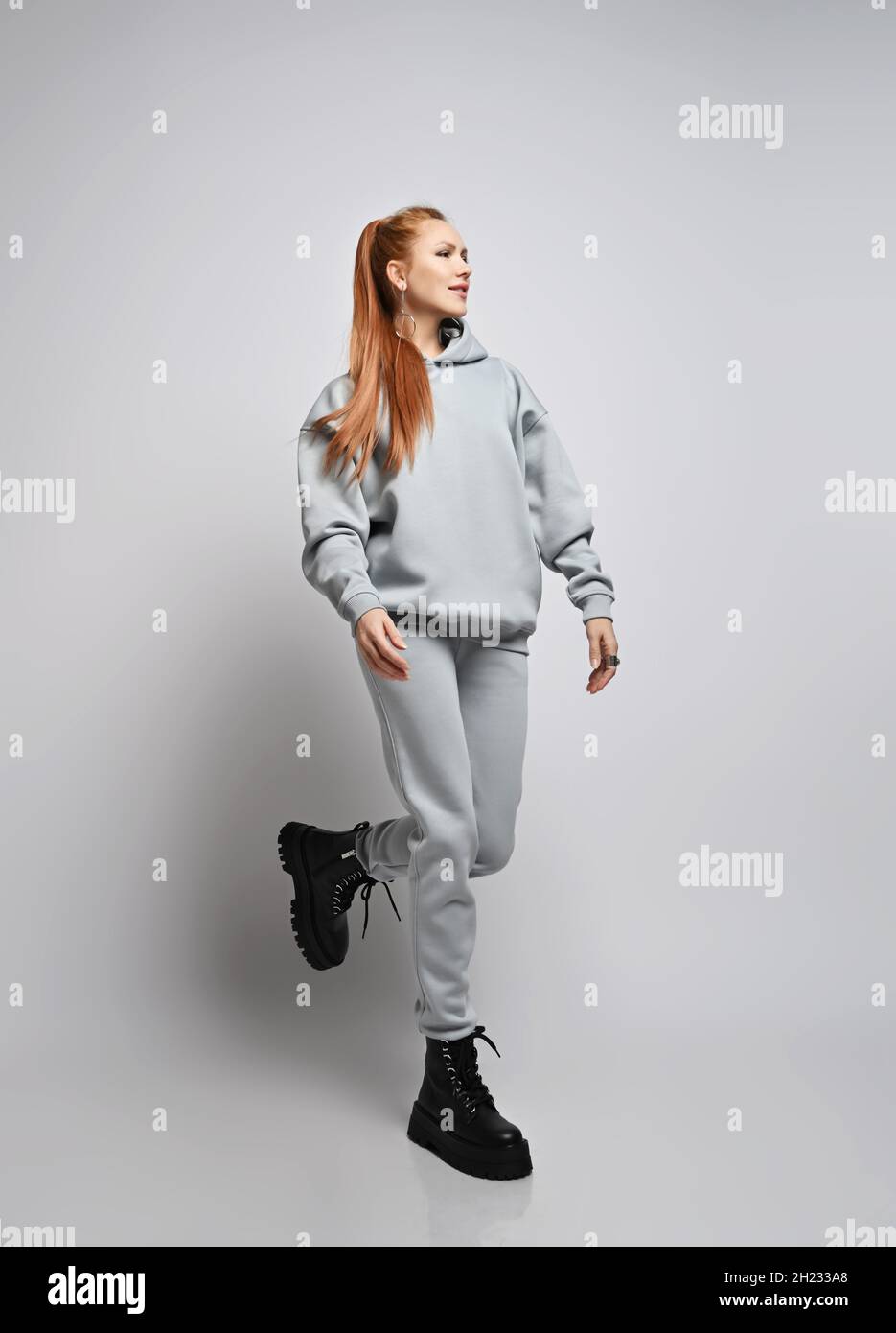 Happy slim red-haired woman with ponytail hairstyle in trendy grey sportswear hoodie, pants and black shoes walks, runs Stock Photo
