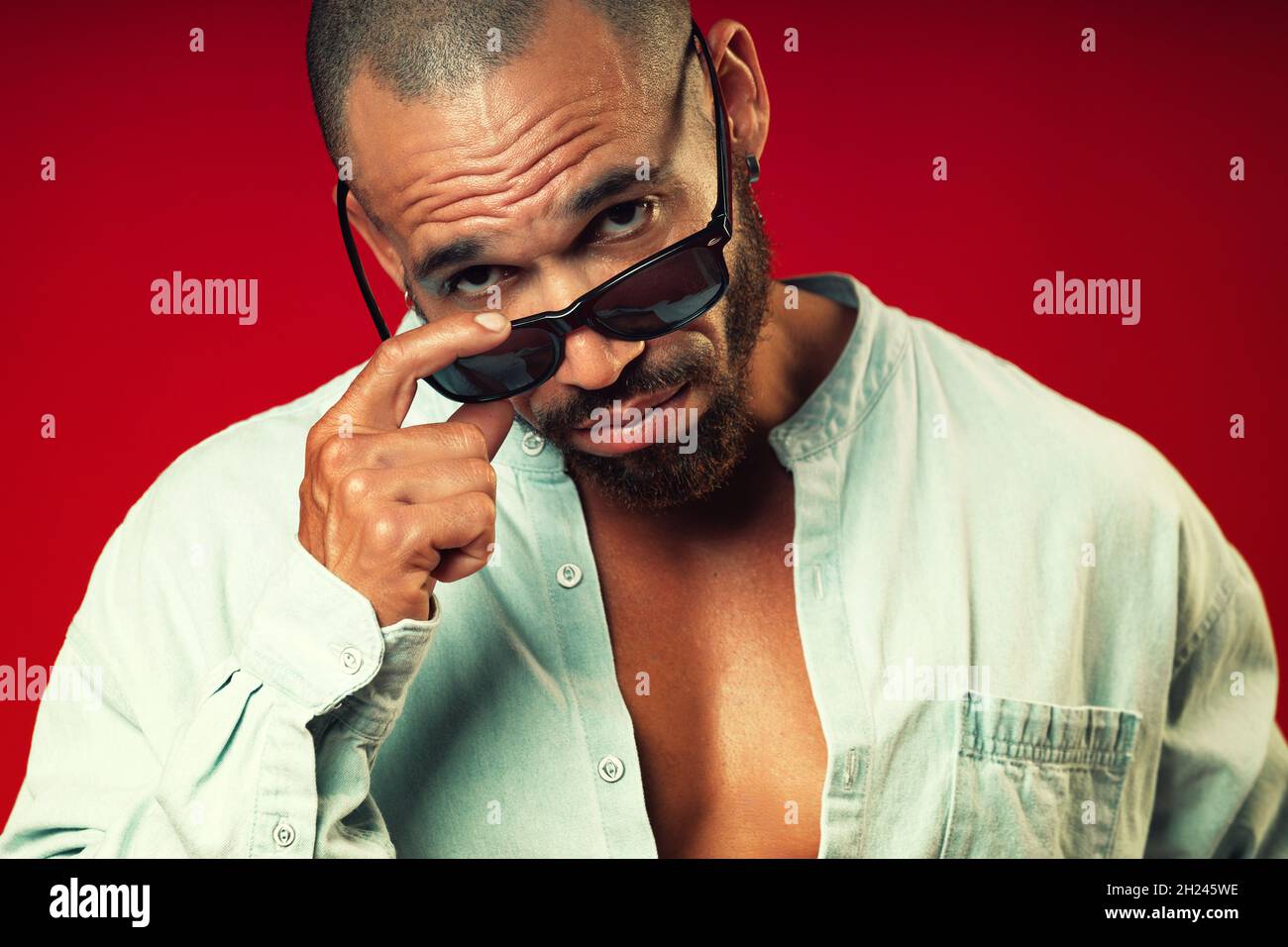 Portrait of a swarthy Latino questioning look from under sunglasses on a red background. Brutal bearded man with a short haircut. Stock Photo