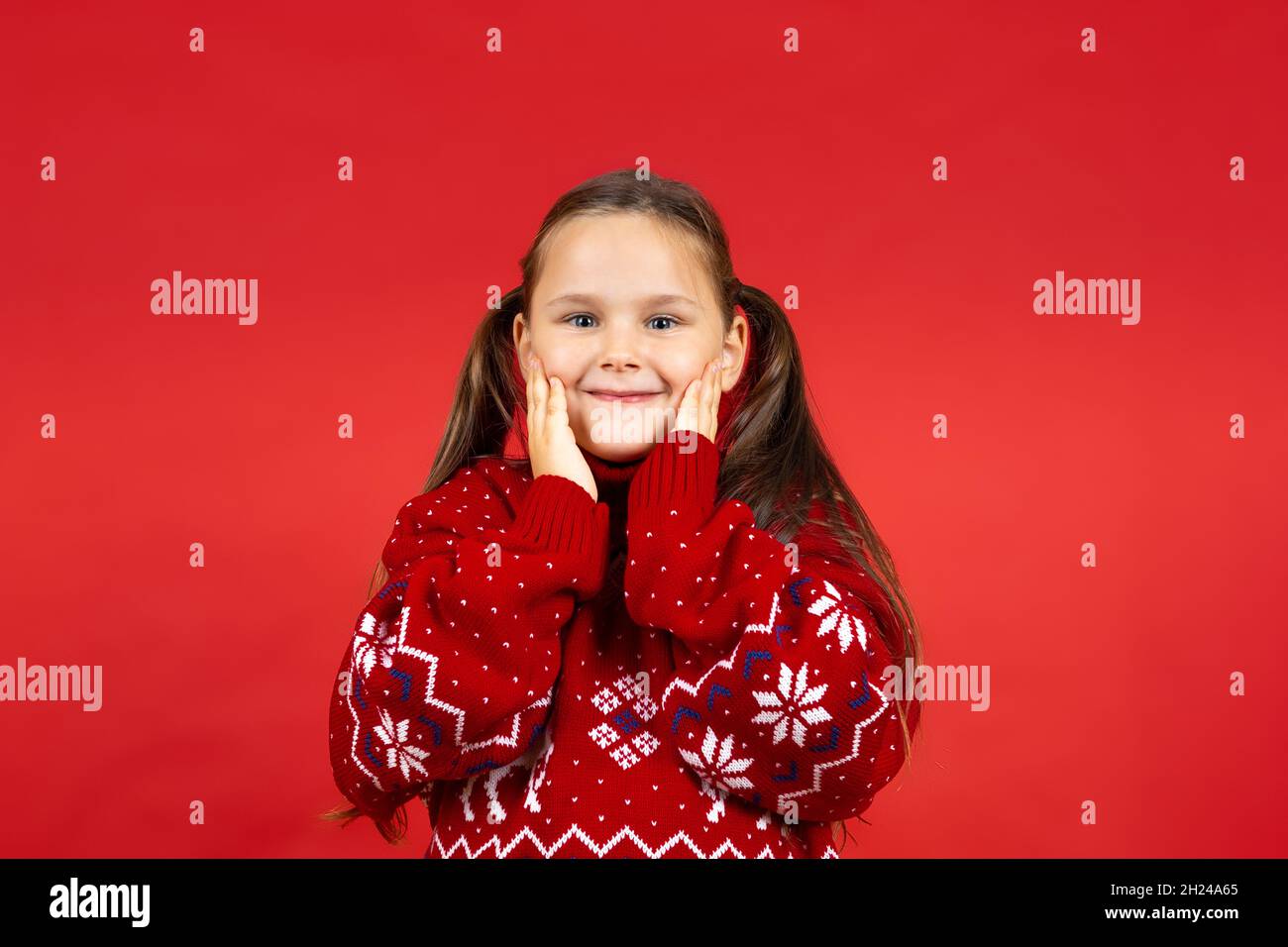 close-up portrait of beautiful happy girl in red knitted Christmas sweater with reindeer touching her face with hands, isolated on red background Stock Photo