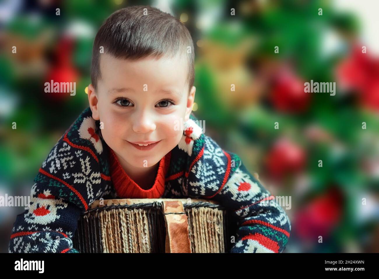 A beautiful happy child in a Christmas sweater smiles and looks at the camera. Caucasian kid on the background of a Christmas tree. Stock Photo