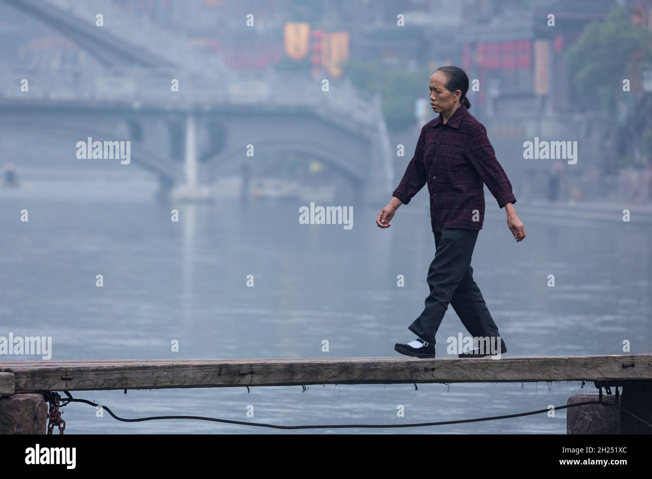 An older Chinese woman walks across a wooden bridge over the Tuojiang River in Fenghuang, China. Stock Photo