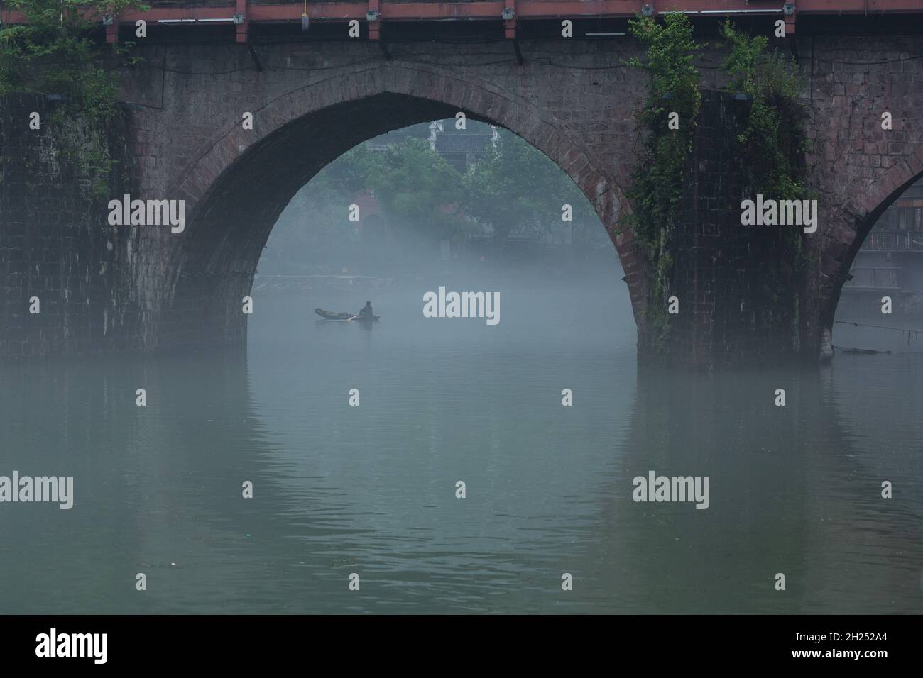 A bridge arch frames a man in a sampan in early-morning fog on the Tuojiang River in Fenghuang, China. Stock Photo