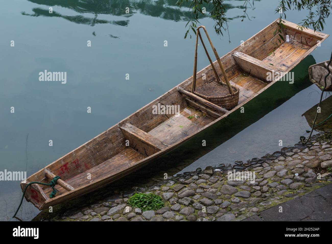 A traditional sampan tied up on  the Tuojiang River with a wicker basket scoop for cleaning the river. Fenghuang, China. Stock Photo