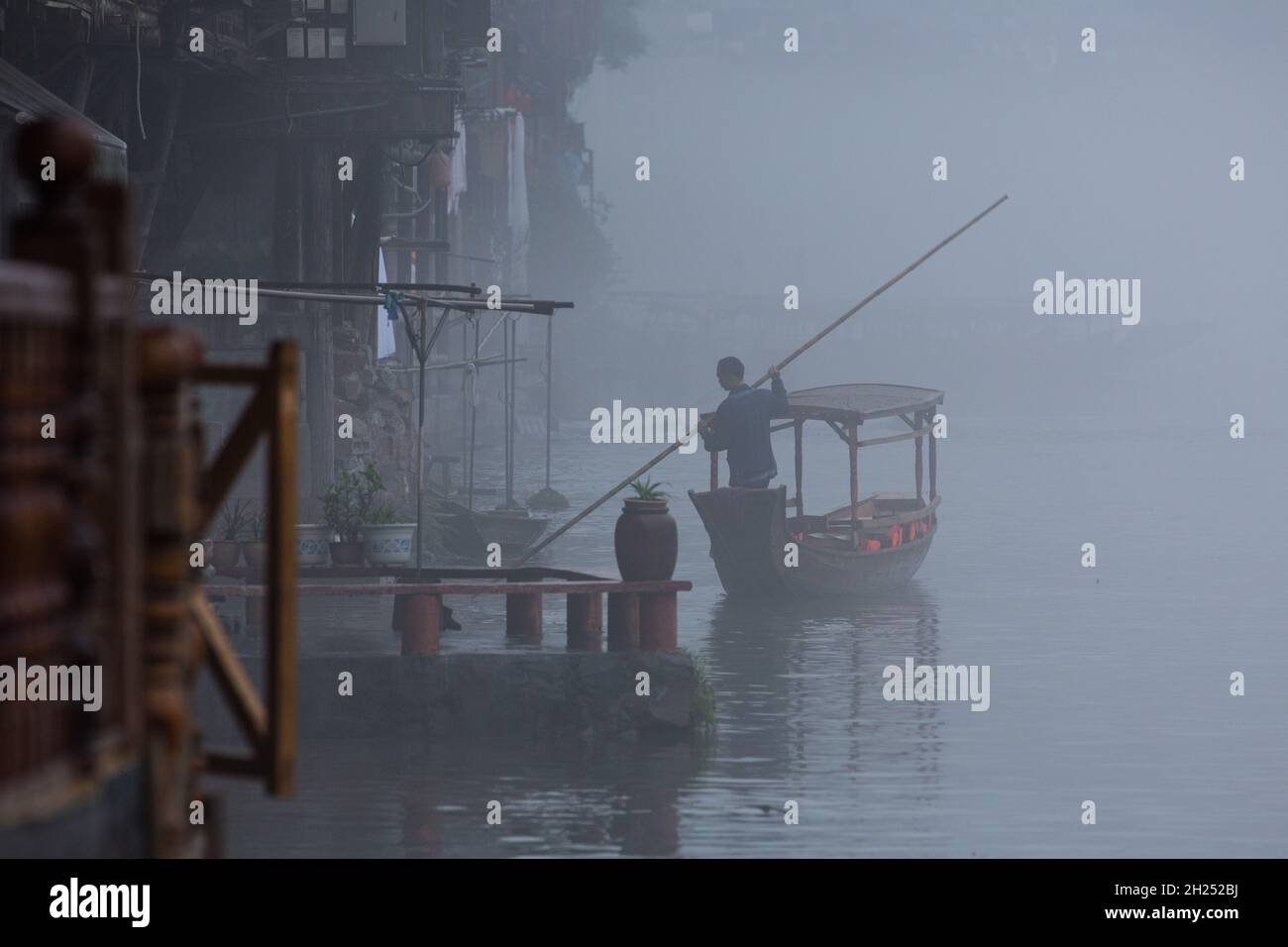 A man poles an empty covered tour boat up the Tuojiang River in early-morning fog.  Fenghuang, China. Stock Photo
