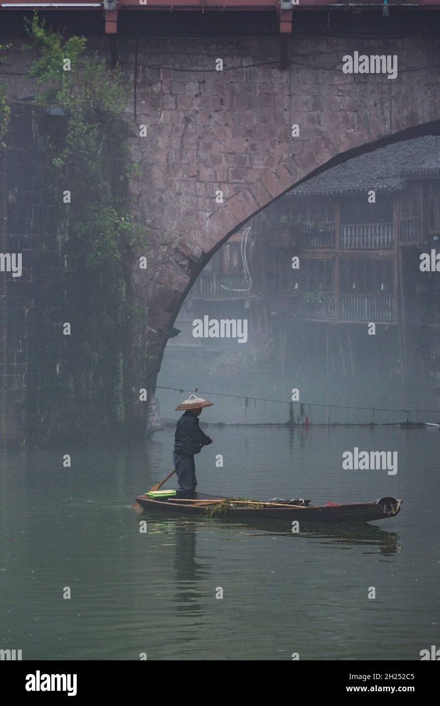 A man paddles a sampan in the fog by the Phoenix Hong Bridge on the Tuojiang River, Fenghuang, China. Stock Photo