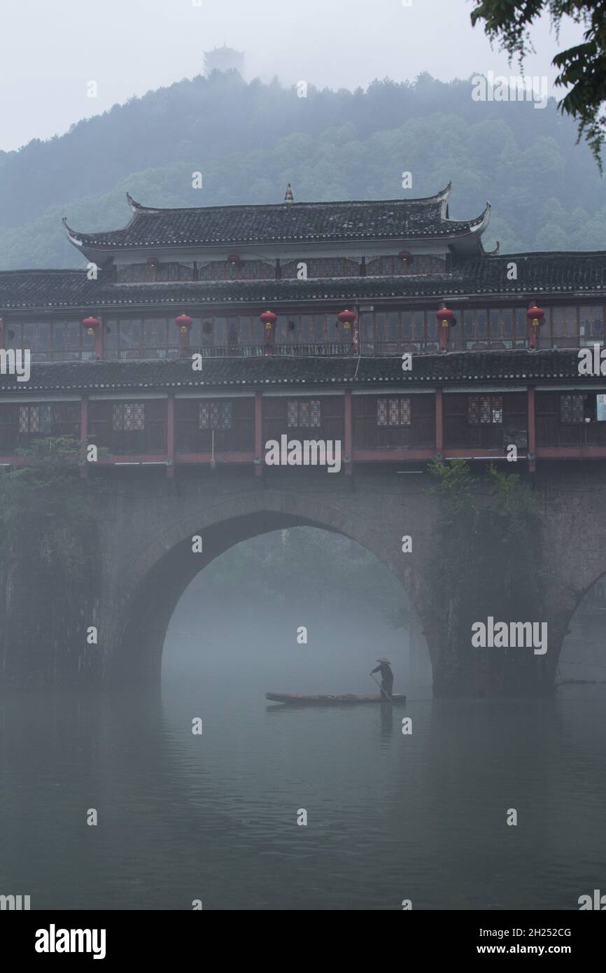 A man paddles a sampan in the fog by the Phoenix Hong Bridge on the Tuojiang River, Fenghuang, China. Stock Photo