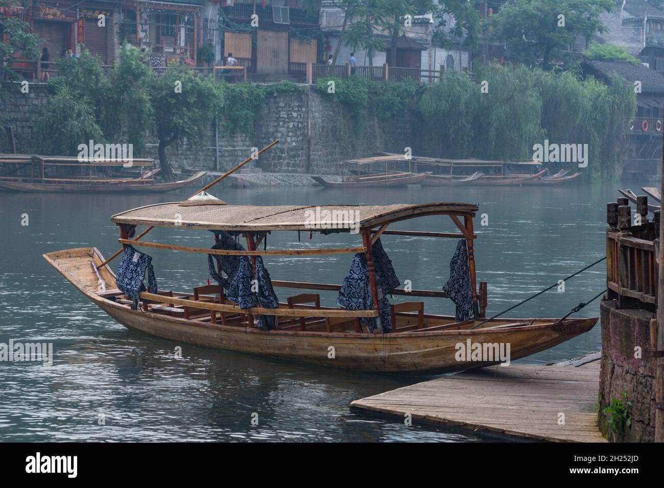 A covered tour boat docked on the Tuojiang River in Fenghuang, China, with a traditional conical hat on top. Stock Photo