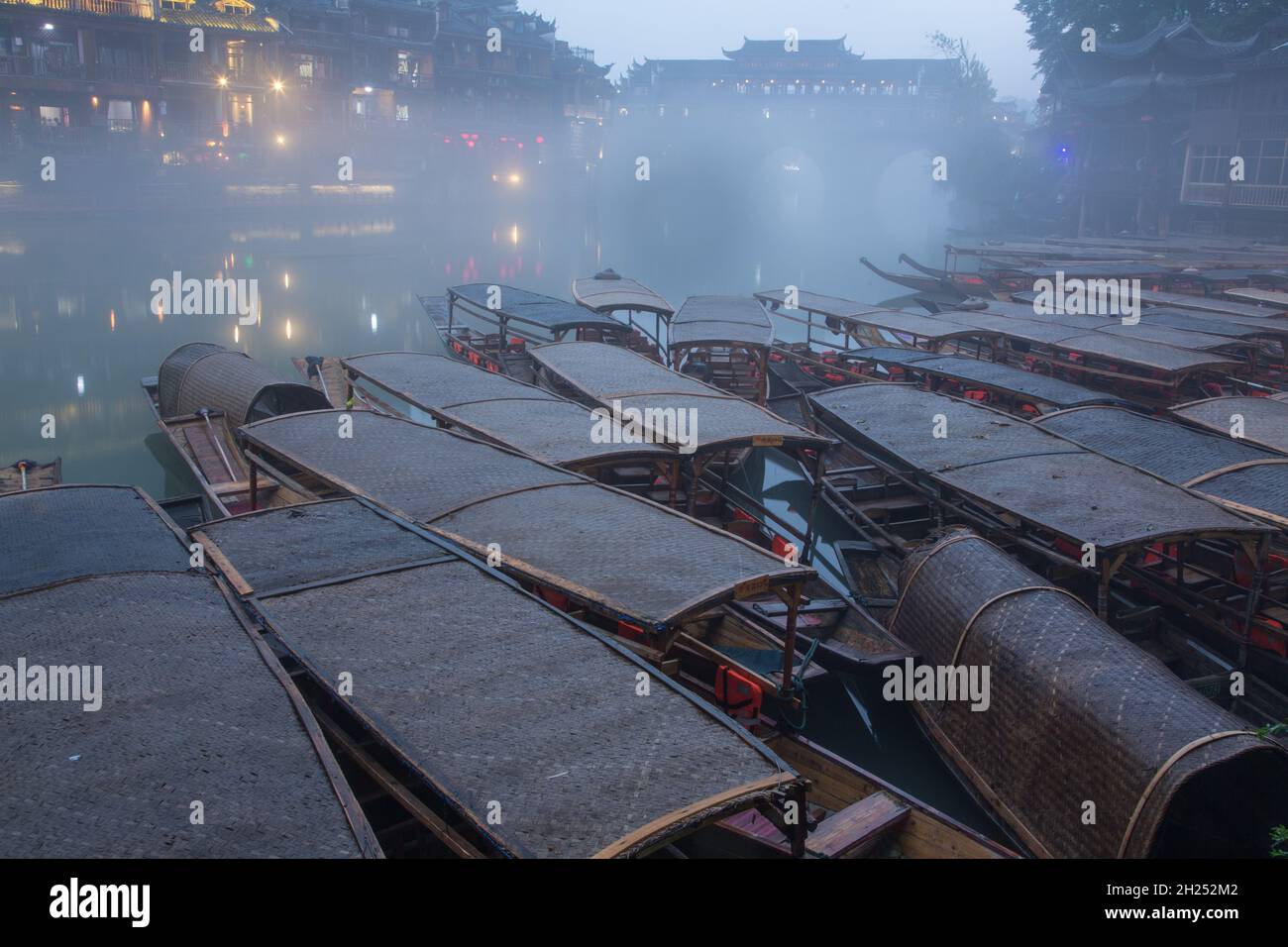Covered tour boats docked on the bank of the Tuojiang River.  The Phoenix Hong Bridge is in fog.  Fenghuang, China. Stock Photo