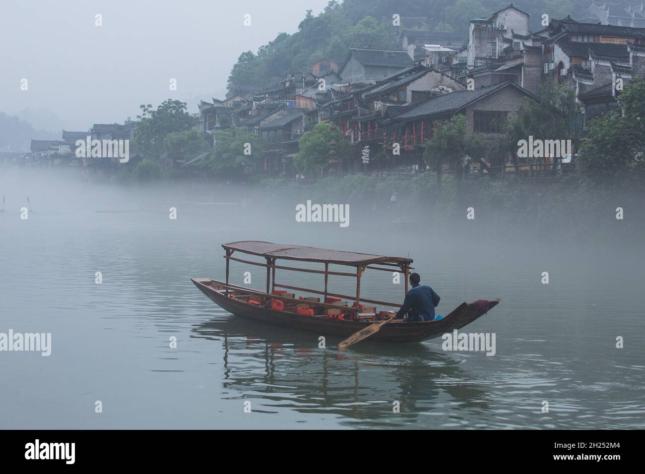 A man paddles a covered tour boat up the Tuojiang River in early-morning fog.  Fenghuang, China. Stock Photo