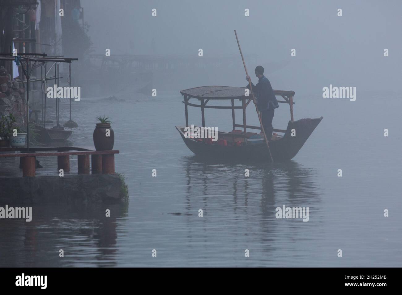 A man poles an empty covered tour boat up the Tuojiang River in early-morning fog.  Fenghuang, China. Stock Photo