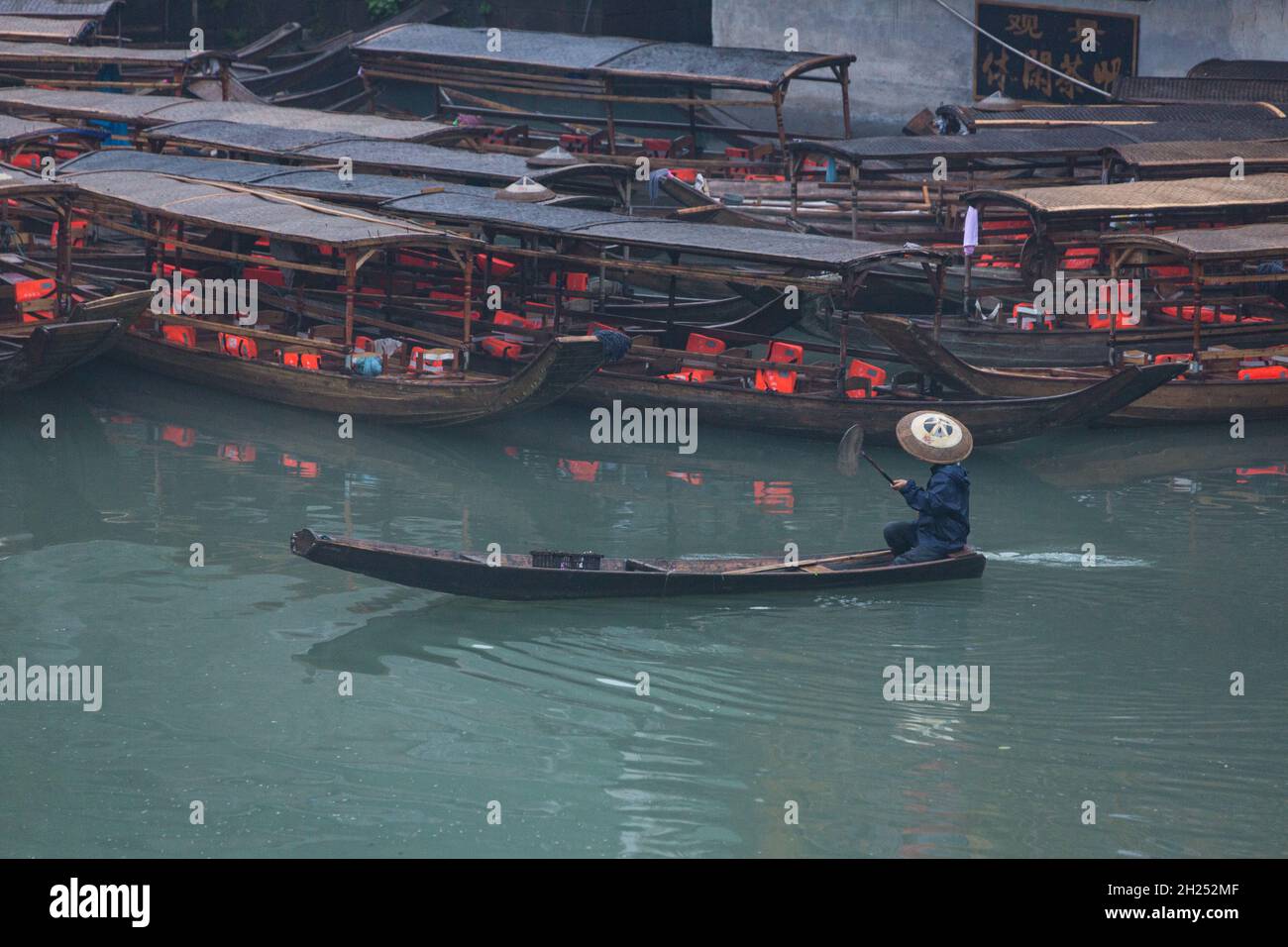 A man in a traditional conical hat paddles a sampan on the Tuojiang River in Fenghuang, China. Stock Photo