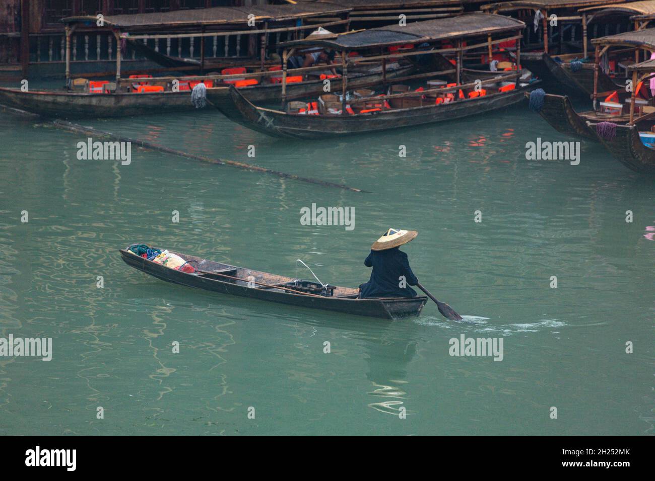 A man in a traditional conical hat paddles a sampan on the Tuojiang River in Fenghuang, China. Stock Photo