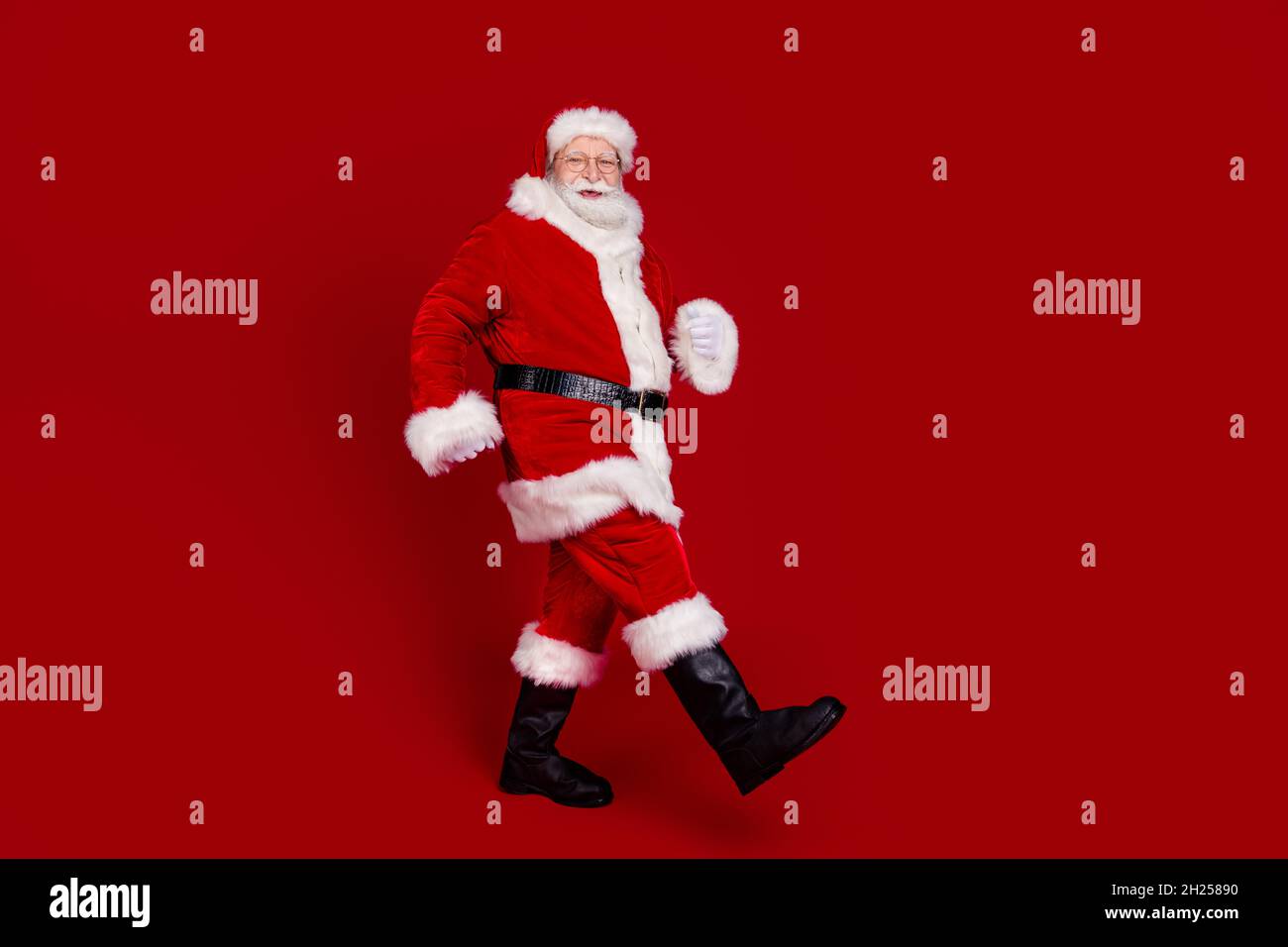 Photo of cute funky man pensioner dressed santa claus costume boots walking empty space smiling isolated red color background Stock Photo