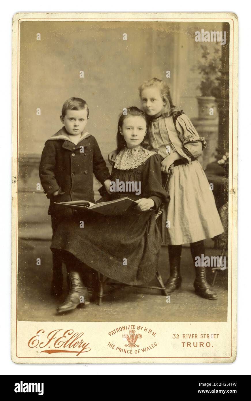 Victorian cabinet card of 3 attractive young Cornish children - not from a wealthy family as the older girl's dress is torn - these are probably their 'Sunday Best' clothes. Photographed by E.I. Ellery, Truro, Cornwall, U.K. This photographer states that his studio of patronized by H.R.H. Prince of Wales (later, Edward V11) circa 1900. Stock Photo