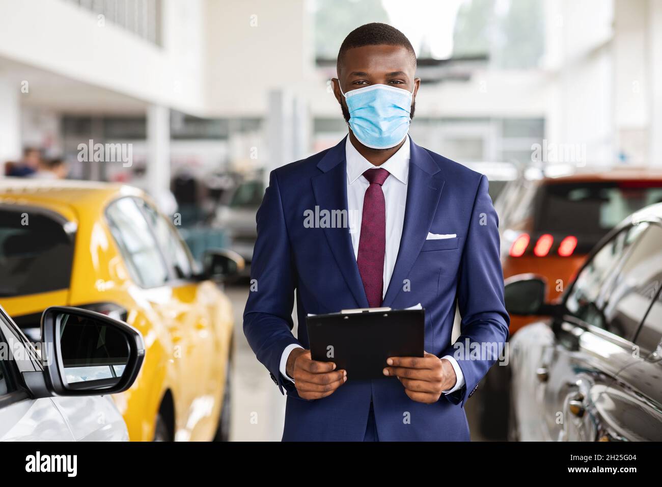 Young Black Car Salesman Wearing Medical Mask Posing At Workplace In Showroom Stock Photo