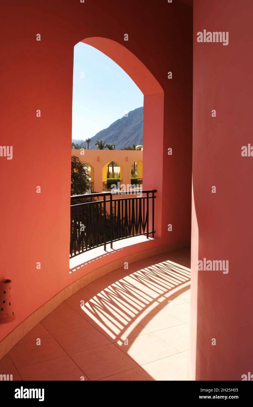 Balcony walkway red walls with  opening to view of sky & hill reflections on floor of railings traditional Egyptian architecture palm trees copy space Stock Photo