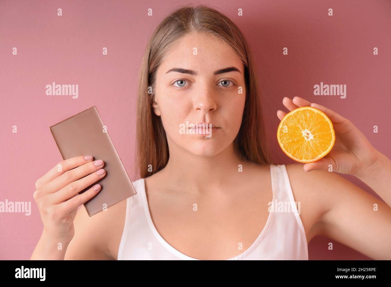 Young woman with acne problem holding orange and chocolate bar on color