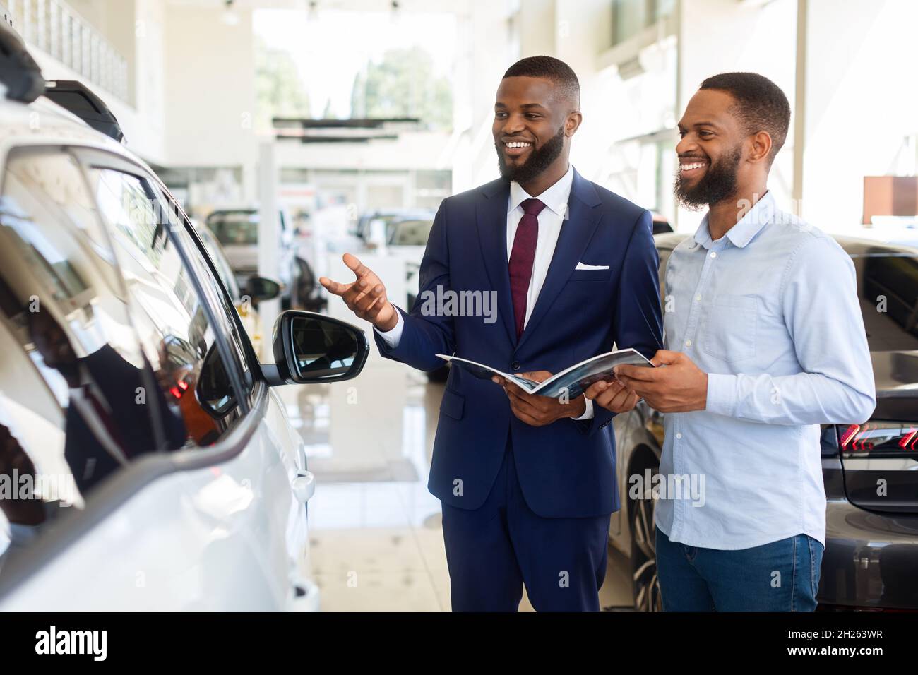Black Salesman Advertising New Automobile To Male Customer In Car Dealership Center Stock Photo