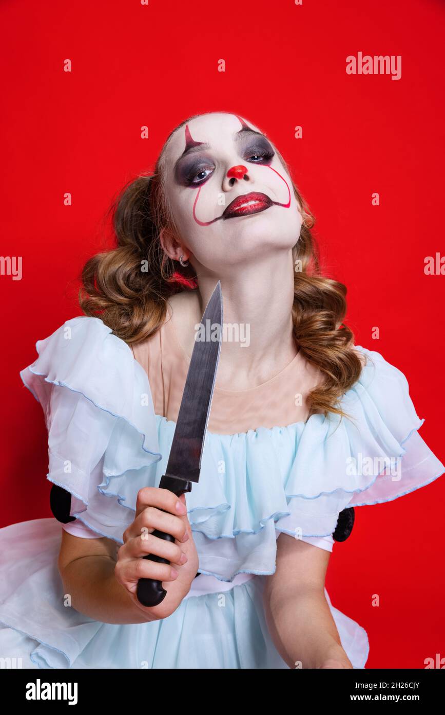 Halloween spooky theme. Mystical blonde girl with bright holiday make up holding knife isolated over red background Stock Photo