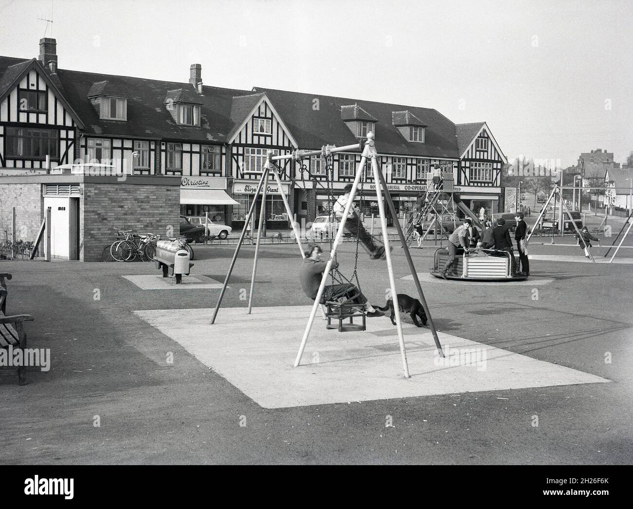 1960s, historical, a view across a childrens playground with swings and roundabout and traditional rocking horse located beside a suburban mock-tudor parade of shops, South London, England, UK, possibly Abbey Wood or Eltham. One of the shops is a Royal Arsenal Co-Operative, at this time a large consumer co-operative trading in the South East London. Stock Photo