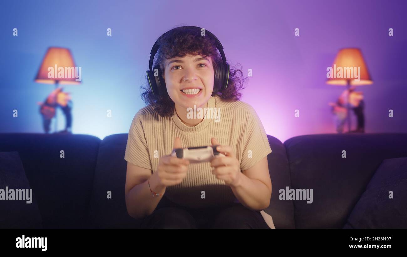 Portrait of excited young gamer woman sitting on couch, playing a video game with wireless controller on console at home. Young gamer girl wearing hea Stock Photo
