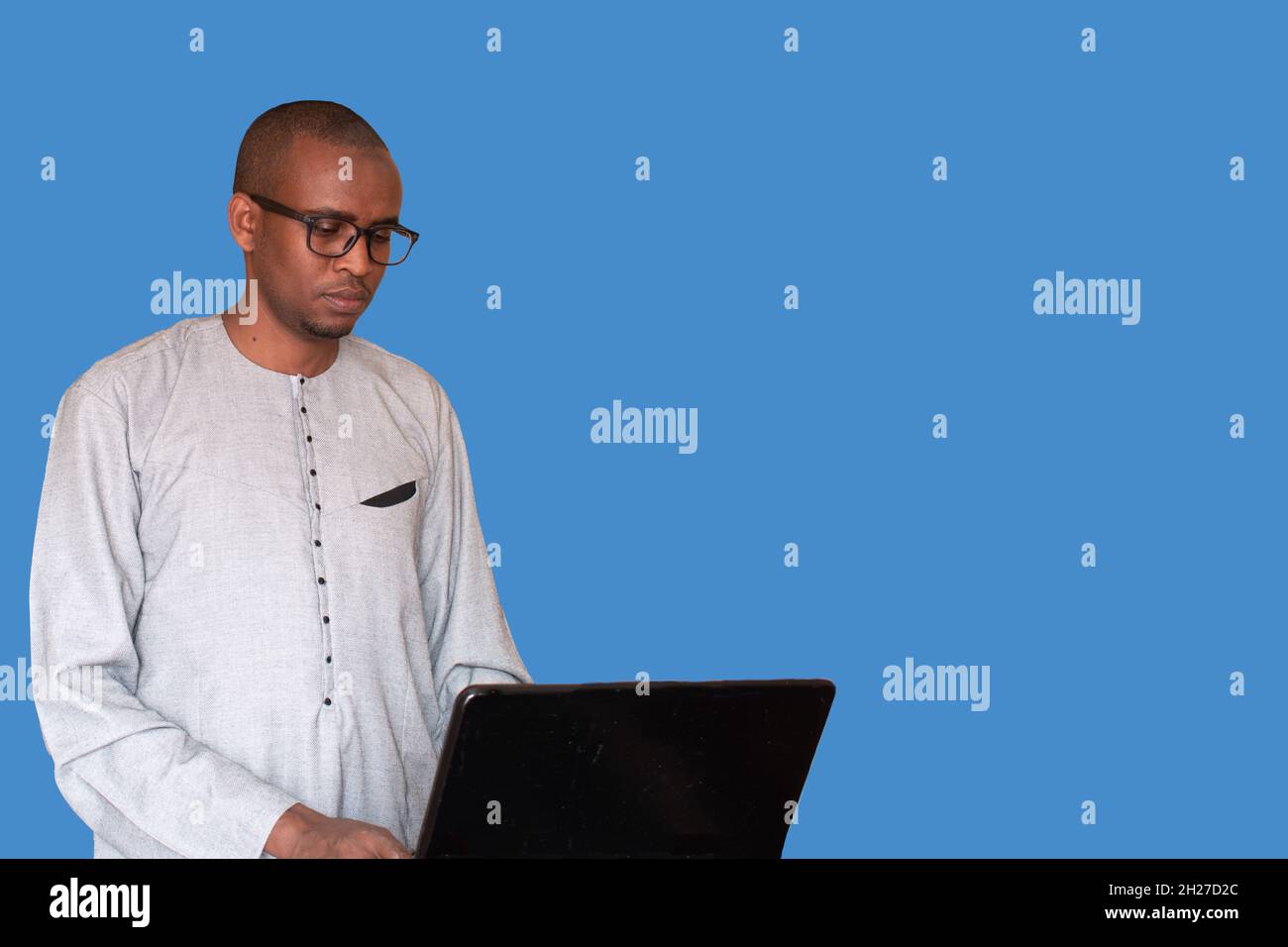 A Muslim man dressed in a kaftan uses his laptop in front of a blue background Stock Photo