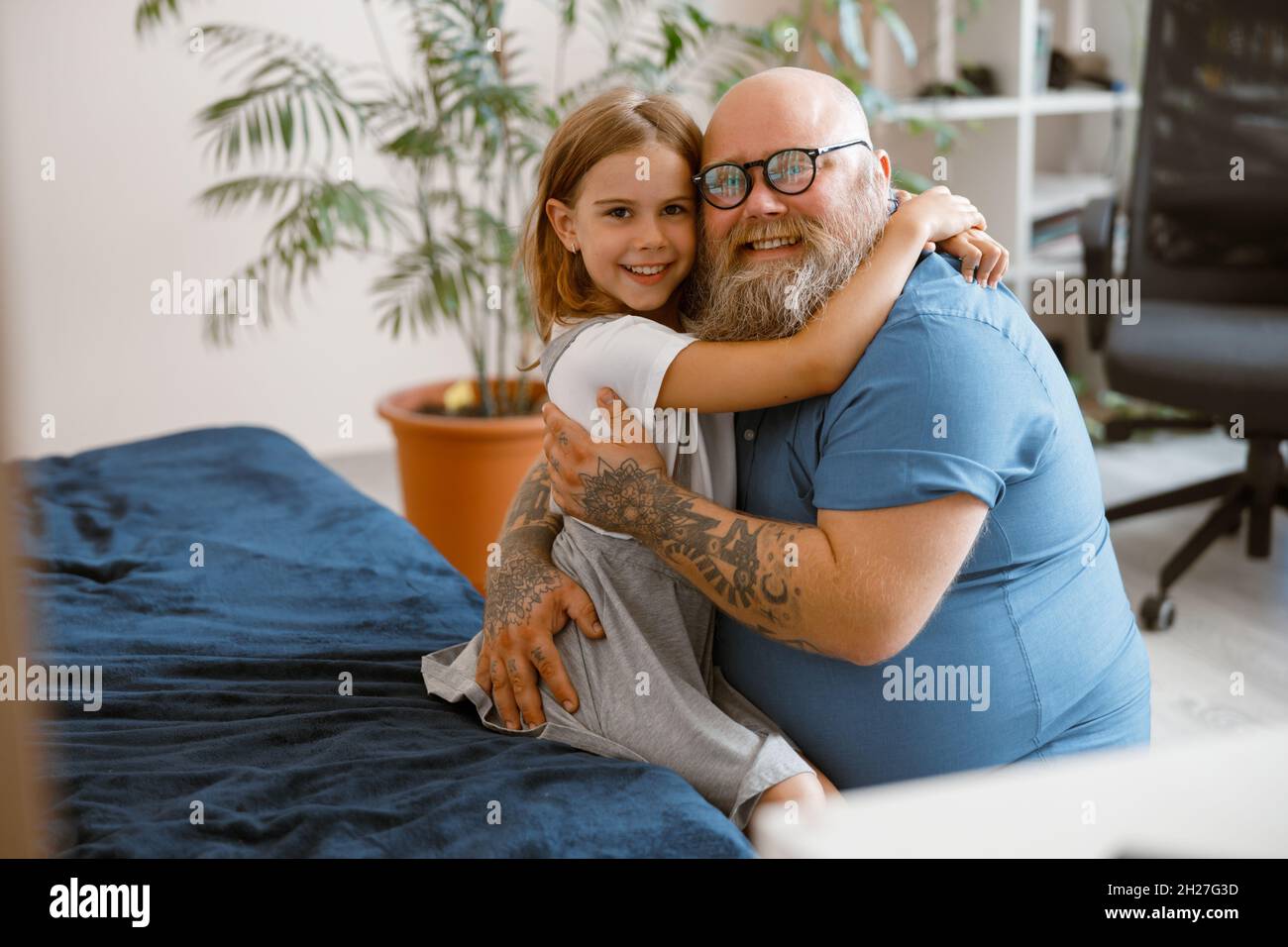 Smiling father with glasses embraces pretty daughter sitting on bed at home Stock Photo