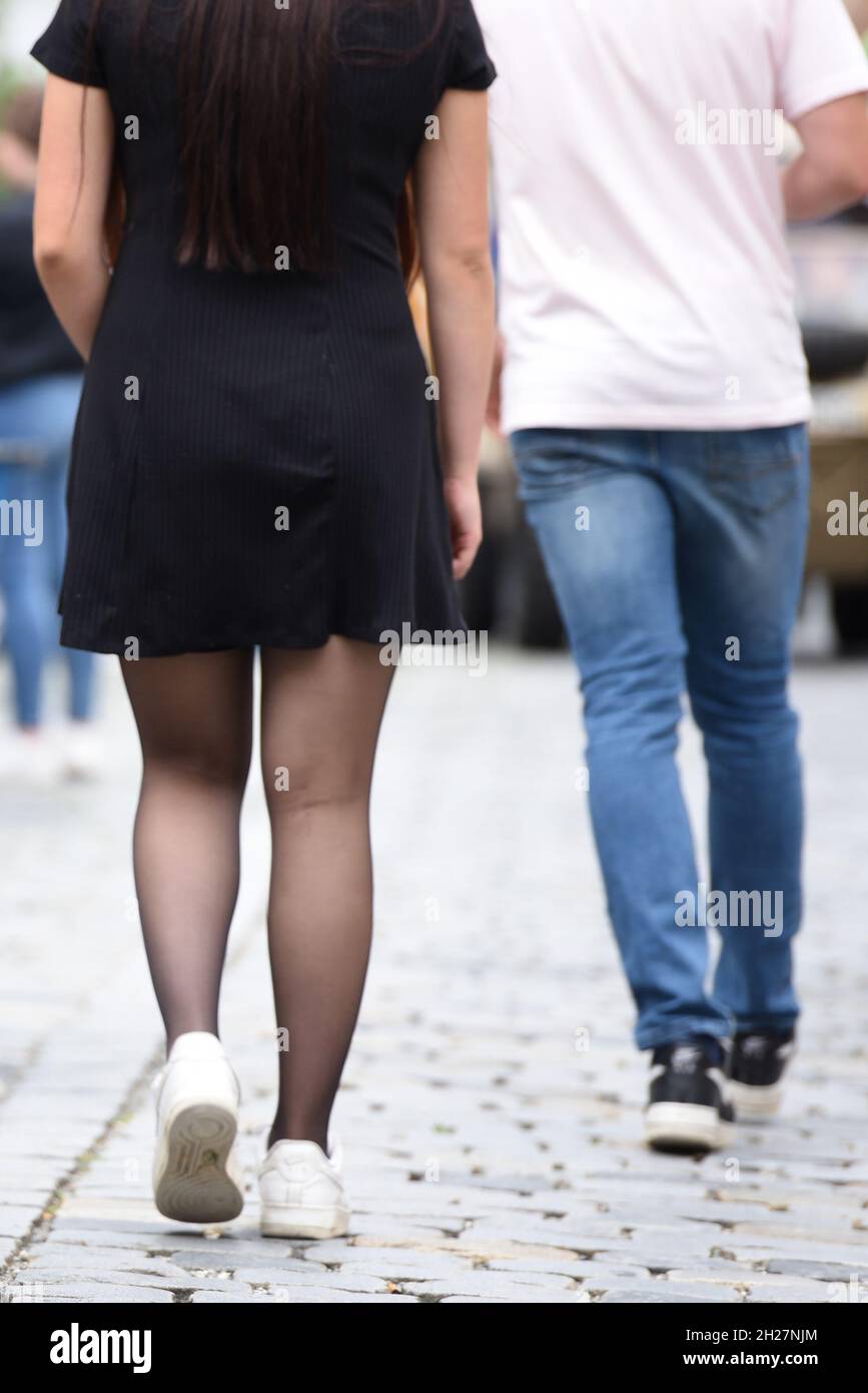 Junge Frau mit schwarzem Minikleid und schwarzen Strünpfen geht hinter einem Mann in Jeans und weißem T-Shirt - Young woman with black mini dress and Stock Photo