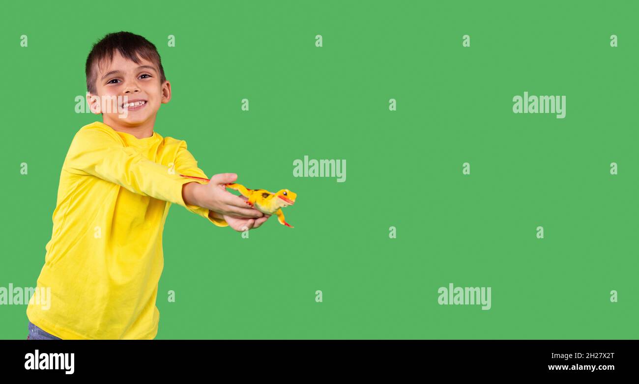 Clever kid boy in yellow hoodie with smile on his face lending his hand with dinosaur toy, standing against a green background studio, copy space. Stock Photo