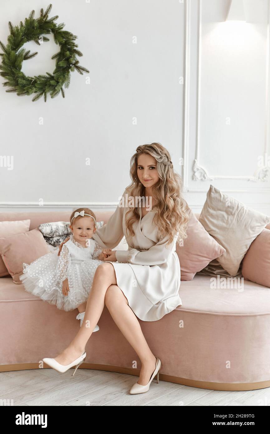 Gorgeous young woman in stylish dress posing with her little daughter in fluffy dress in Christmas decorated interior Stock Photo