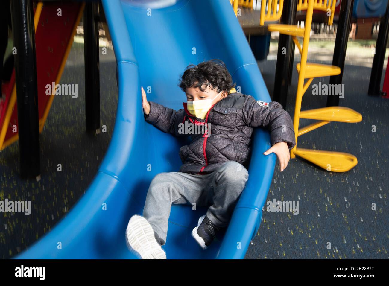 Education Preschool 4-5 year olds playground boy sliding down slide, wearing face mask to protect against Covid-19 infection Stock Photo